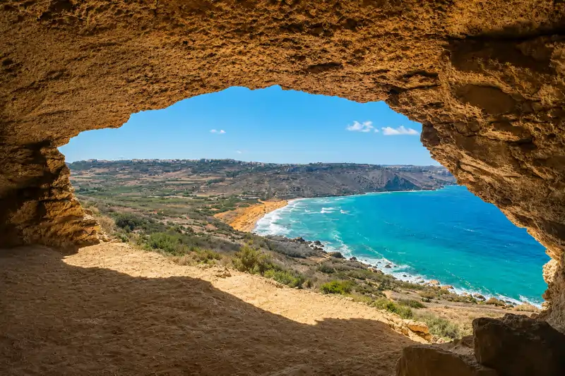 A turquoise bay and terraced hillsides seen through the mouth of a limestone cave on the Gozitan coast.